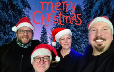 Vier Personen mit Weihnachtsmützen vor einem weihnachtlichen Hintergrund. Über ihnen steht die Aufschrift Merry Christmas.