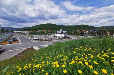 Im Vordergrund ist eine Wiese mit gelben Blumen. Dahinter befindet sich ein Parkplatz. In der Ferne sieht man Häuser und Wälder.