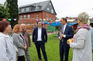 Eine Gruppe Männer und Frauen steht im Halbkreis auf einer Wiese vor einem Backsteinhaus.