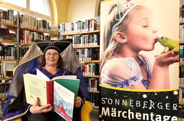 Ein Frau sitzt neben einem Banner mit der Aufschrift: Sonneberger Märchentage. Sie hält ein Buch in der Hand.