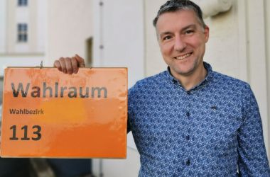 Ein Mann hält ein oranges Schild mit der Aufschrift: Wahlraum Wahlbezirk 113.