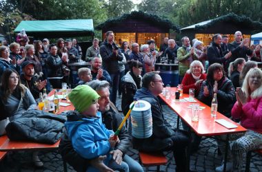 Menschen sitzen an Bierzeltgarnituren. Dahinter stehen viele weitere Personen. Im Hintergrund stehen Verkaufsstände.