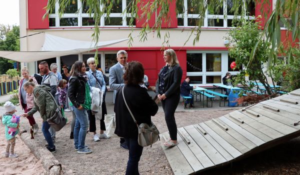 Kinder und Erwachsene stehen auf einem Spielplatz vor einem roten Gebäude.