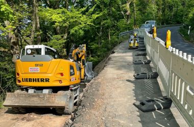 Ein Bagger steht links neben dem Straßenrand. Auf der Straße steht eine Absperrung.