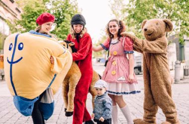 Vier Personen in Spielzeugkostümen stehen in einer Reihe und halten sich an den Schultern. Vor ihnen steht ein kleines Kind.