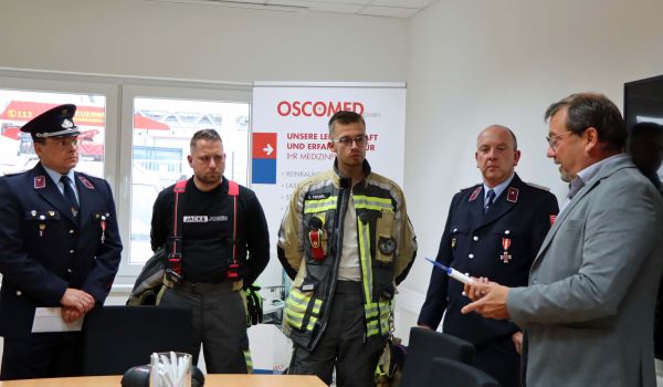 Vier Feuerwehrmänner stehen in einem Büro und bekommen von einem Mann ein medizinisches Gerät erklärt.