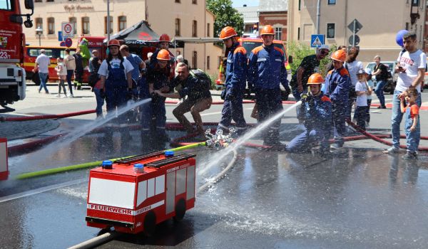 Kinder in Feuerwehr-Uniform zielen mit Wasserschläuchen auf kleine Feuerwehrautos.