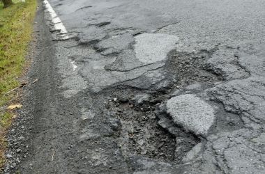 Eine Straße mit Schlagloch. Der Asphalt ist abgebröckelt. Im Hindergrund kommt ein blauer LKW angefahren.