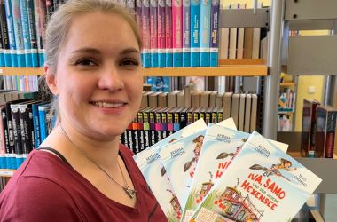Eine Frau steht vor einem Bücherregal. Sie hält Kinderbücher mit der Aufschrift - Iva, Samo und der geheime Hexensee - hoch.