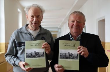 Zwei Männer halten ein Buch mit der Aufschrift: Landwirtschaft Hönbach in den Händen. Darunter ist ein altes Schwarz-Weiß-Foto.