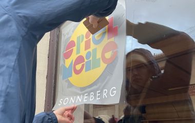 Eine Frau klebt ein Schild an eine Glasscheibe mit der Aufschrift: Spiel Meile Sonneberg.