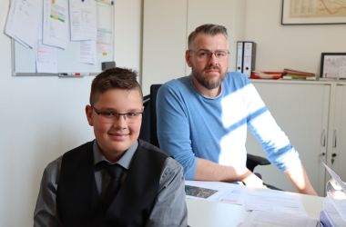 Ein Mann sitzt an einem Schreibtisch. Vor dem Tisch sitzt ein Junge.