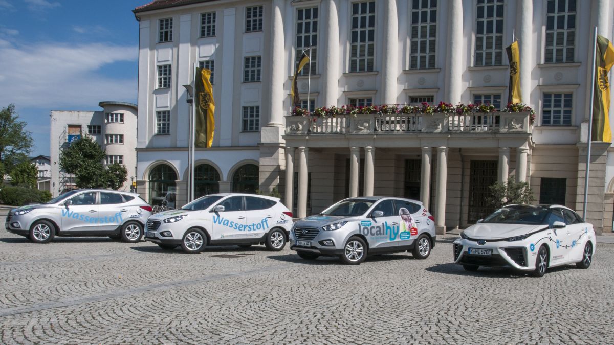 Vier Wasserstoff-Autos stehen auf einem Platz. Dahinter befindet sich das Rathaus der Stadt Sonneberg.