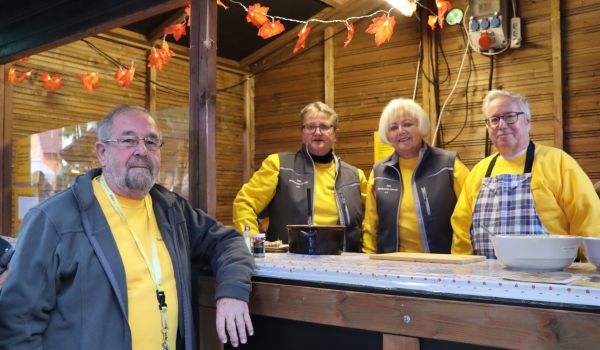 Drei Personen stehen hinder einem Verkaufsstand aus Holz. Ein weiterer Mann steht davor.