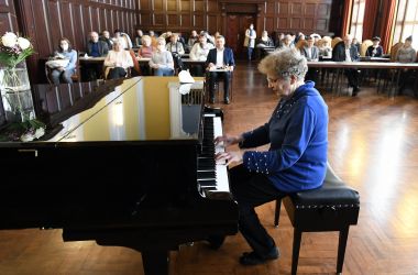 Eine Frau sitzt vor einem Flügel und spielt darauf. Im Hintergrund sitzen weitere Personen.
