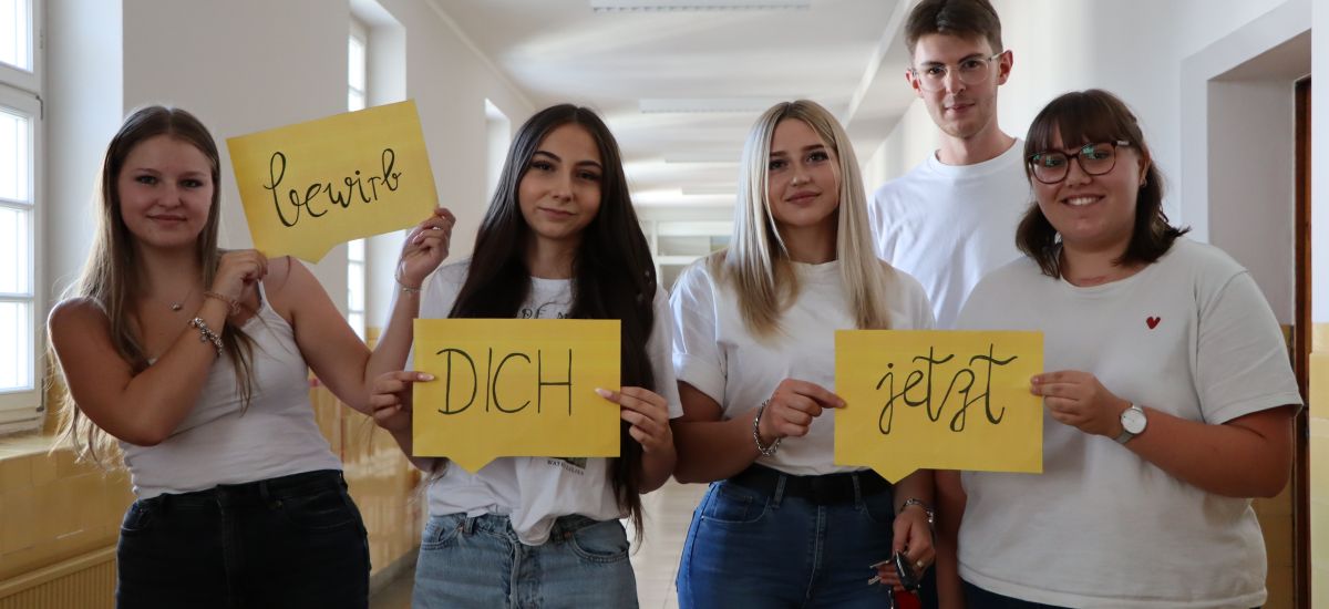 Fünf junge Menschen stehen in einem Flur. Drei von ihnen halten Schilder mit den Aufschriften: Bewirb - dich - jetzt.