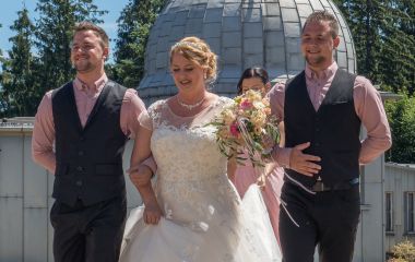 Eine Frau im Hochzeitskleid wird von zwei Männern zum Altar geführt. Im Hintergrund ist eine Kuppel der Sternwarte Sonneberg zu sehen.