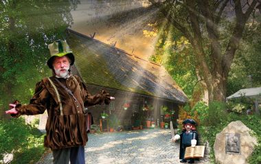 Ein als Spielmann verkleideter Mann steht vor einem alten Holzhaus.