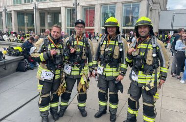 Vier Feuerwehrmänner in Schutzkleidung stehen nebeneinander auf einem großen Platz mit vielen Menschen. Sie tragen eine Medaille um den Hals.