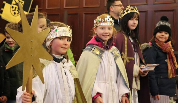Kinder verkleidet als Sternsinger stehen vor einer Holzwand. Sie tragen goldene Kronen und lange Mäntel.