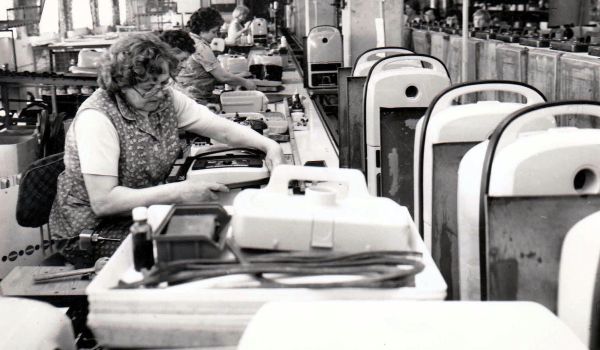 Schwarz-weiß: Frauen sitzen nebeneinader an Tischen in einer Fabrik und fertigen Staubsauger.