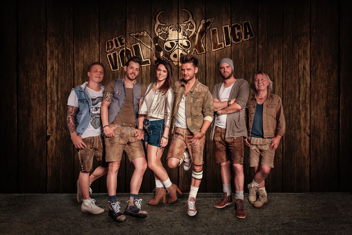 Eine Volksmusikgruppe in Lederhosen posiert vor einer Holzwand mit der Aufschrift: Die VolXXLiga.