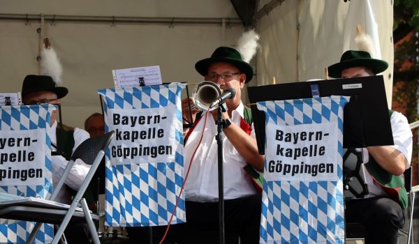 Nahaufnahme: Männer in Tracht sitzen auf einer Bühne und spielen Blechblasinstrumente. Auf ihren blau-weiß-karierten Notenständern steht die Aufschrift: Bayernkapelle Göppingen.
