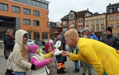 Ein Mann mit gelber Jacke übergibt ein Luftballontier an ein Kind.