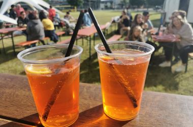 Zwei Becher mit orangen Cocktails stehen auf einem Tisch. Im Hintergrund sitzen Menschen auf Bierbänken. Es scheint die Sonne.