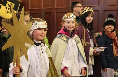 Kinder verkleidet als Sternsinger stehen vor einer Holzwand. Sie tragen goldene Kronen und lange Mäntel.