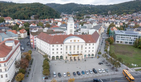 In der Mitte steht ein weißes Gebäude mit einem roten Dach und einem Turm. Es ist das Rathaus der Stadt Sonneberg. Rechts davon ist eine Wiese. Auf der linken Seite steht das AOK-Gebäude. Im Hintergrund ist die Stadt.