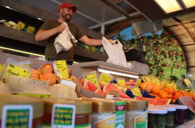 Auf einem Marktstand liegt frisches Obst. Im Hintergrund steht ein Verkäufer.