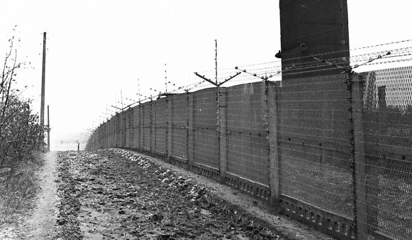 Schwarz-weiß: Ein Grenzzaun mit einem Wachturm daneben.