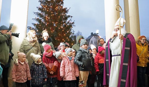 Ein Mann in Nikolaus-Kostüm mit goldenem Zepter. Um ihn herum stehen viele Kinder. Im Hintergrund steht ein großer Weihnachtsbaum.