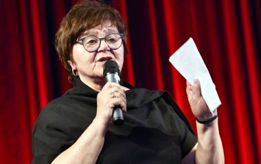 Eine Frau steht auf einer Bühne und hält ein Mikrofon und einen Zettel in der Hand.