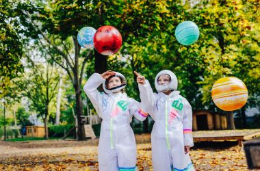 Zwei Kinder in weißen Astronautenanzügen stehen auf einem Spielplatz. Über ihnen hängen Planeten.