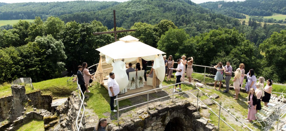 Ein Hochzeitspavillon steht auf einer Burgruine. Außenrum stehen Gäste.