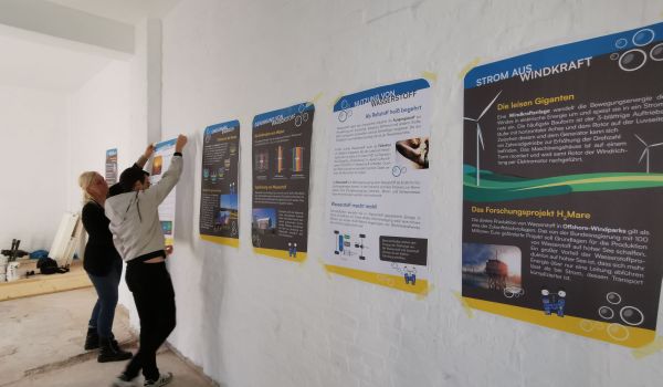Zwei Personen kleben Plakate mit Informationen zu Wasserstoff an eine Wand.