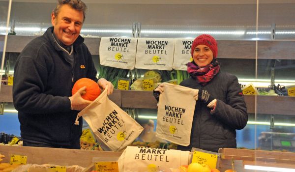 Zwei Markthändler packen Gemüse in einen Stoffbeutel mit der Aufschrift: Wochenmarktbeutel.
