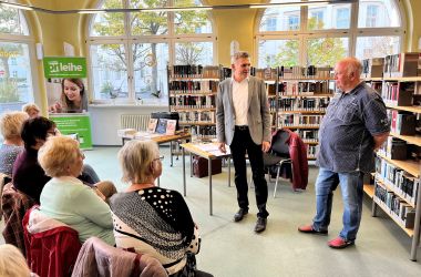 Links sitzen Frauen in Stuhlreihen. Davor stehen der Bürgermeister Dr. Heiko Voigt und Harald Saul. Im Hintergrund sind Bücherregale.