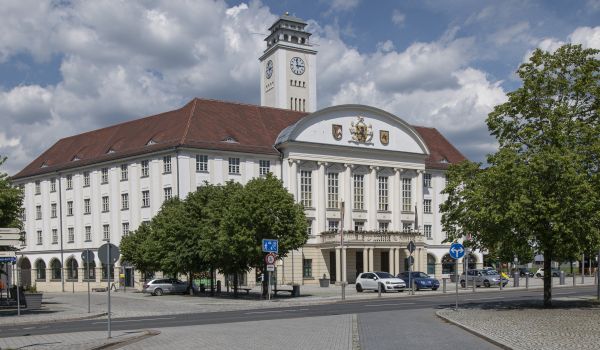 EIn weißes Gebäude mit rotem Dach und einem Turm. Es ist das Rathaus der Stadt Sonneberg. Davor stehen einzelne Bäume Die Sonne scheint.