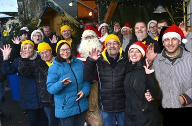 Viele Menschen winken auf einem Weihnachtsmarkt. In der Mitte steht der Weihnachtsmann.