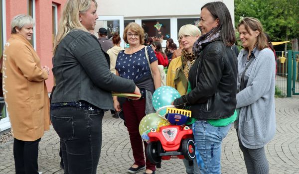 Frauen stehen in einem Kreis vor einem Gebäude. Eine der Frauen hält ein roter Bobby-Car mit Luftballons in der Hand.
