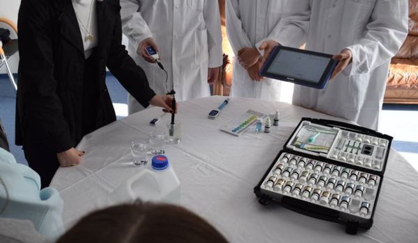 Auf einem Tische liegt ein Experimentierkoffer. Dahinter stehen Menschen in weißen Kitteln.