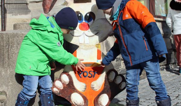 Zwei Kinder werfen Briefe in den Spalt eines Holzaustellers. Der Aufsteller bildet einen Osterhasen ab. Unter dem Spalt steht die Aufschrift: Post.