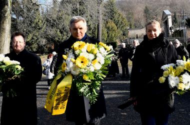 Drei schwarzgekleidete Personen bringen Blumen auf einen Friedhof.