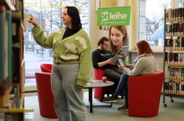 Im Vordergrund steht eine Frau und nimmt ein Buch aus einem Regal. Im Hintergrund sitzen zwei Personen in roten Sesseln und lesen.
