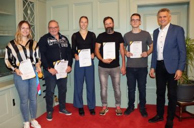 Fünf Personen stehen in einer Reihe nebeneinander. Sie halten eine Urkunde in den Händen. Rechts daneben steht der Bürgermeister.