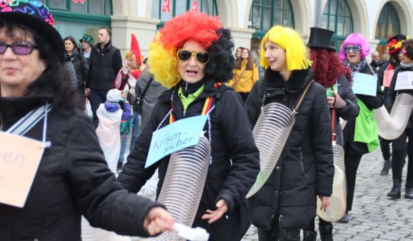 Schwarzgekleidete Personen mit bunten Perücken sind durch Plastikröhren miteinander verbunden.
