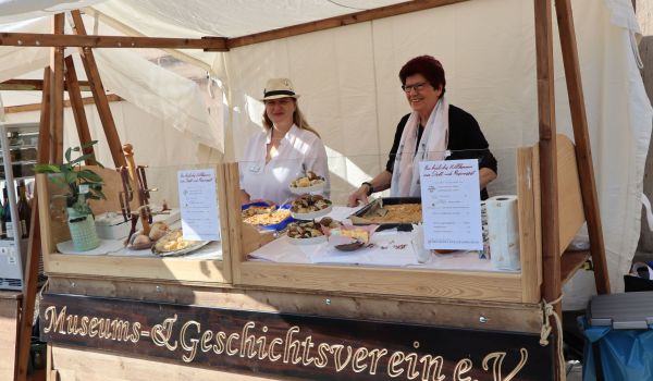Zwei Frauen stehen hinter einem Verkaufstand aus Holz. Auf der Verkaufstheke stehen verschiedene Kuchen. Vorne am Stand ist ein Schild mit der Aufschrift: Museums- und Geschichtsverein e.V..